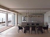Dark dining table and matching chairs on wooden floor; kitchen counter and designer pendant lamps in open-plan interior with view of sea across terrace