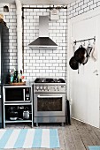 Stainless steel gas cooker below extractor hood against white-tiled wall and blue and white stripes rugs on rustic wooden floor