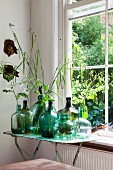 Collection of demijohns in various shades of green, some holding leaves, on vintage folding table next to lattice window