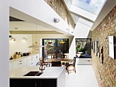 Strip of skylights with white frames and old brick walls in open-plan kitchen with dining area and view of seating area on summery terrace