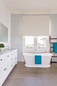 Freestanding country house bathtub in front of window with roller blind next to white vanity unit