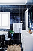 Bathroom with dark wall and floor tiles, corner bath with step and white vanity unit