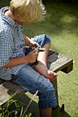 Blonder Junge schnitzt eine Holzpfeife am Holzsteg beim Gartenteich