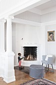 Grey pouffe and armchair in seating area next to fireplace next to white, antique-style column