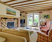 Wood-beamed ceiling above sofa and armchairs around fireplace with firewood store in living room