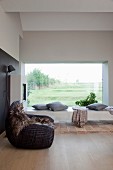 Fur blanked on rattan armchair next to concrete chaise and panoramic window with view of landscape