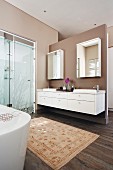 Grosszügiges Bad mit hellbraun getönten Wänden, Teppich vor Waschzeile, seitlich verglaster Duschbereich