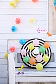 Colourful balloons pinned to dartboard below garlands of paper flowers on white wooden wall