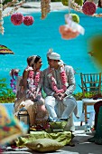 Indian bride and groom adorned with pink flower garlands against backdrop of turquoise sea