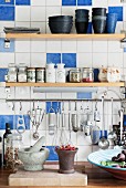 Shelves of spices and crockery above cooking utensils hung from stainless steel rod on kitchen wall with white and blue tiles
