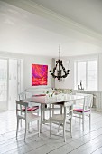 White table and wooden chairs below chandelier in rustic interior with white wooden floor and modern artwork on wall