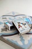 Three matchboxes with pictures on tops arranged on ornamental tile
