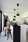 White fitted kitchen with black counter, matching pendant lamps and woman sitting on bar stool