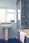 Pedestal sink below window next to bathtub against wall tiled in various shades of blue and red