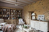 Ländliches Wohnzimmer mit Bücherschrank und gemütlichen Armlehnsesseln, vor rustikal gestalteter Wand weisses Sideboard