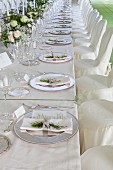 Long festive table with decorated place settings, silver plates, chairs with white covers in front of it