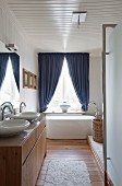 Washstand with wooden base unit and two countertop basins in bathroom with free-standing modern bathtub below window with blue curtains