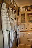 Vintage tea towels and aprons hanging from hooks in front of vintage-style white dresser in corner of kitchen