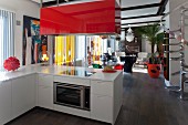 White L-shaped kitchen counter with red-painted extractor hood and lounge area in background