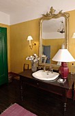 Schreibtisch als Waschtisch mit eingebautem Waschbecken mit Tischleuchte vor Goldrahmenspiegel an gelb getönter Wand