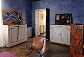 Retro-Stuhl mit Ledergeflecht zwischen weißen Vintage Sideboards in mediterranem Schlafraum mit blau getönter Wandgestaltung und modernen Bildern