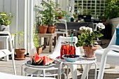 Watermelon, drinks and potted geranium on set of white side tables on terrace