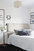 Bed with fabric headboard with ornamental studs, bedside lamp on bistro table and black and white framed pictures