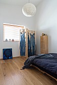 Bed with dark blue bed linen and dungarees hung from wooden screen next to window in minimalist interior