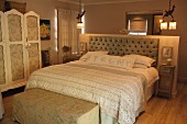 Beige patterned bedspread on double bed with button-tufted headboard against half-height well in rustic bedroom with subdued lighting