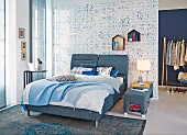 Double bed with denim-upholstered frame in bedroom with blurred patterns on wallpaper and rug
