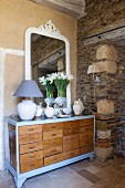 Vase of white flowers and table lamp in front of mirror on top of chest of drawers next to rustic stone wall