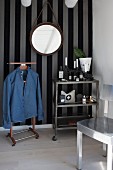 Denim shirt hung on valet stand next to serving trolley below round mirror on striped wallpaper