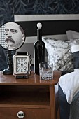 Bottle, glass, alarm clock and vintage table lamp with portrait of man on lampshade on bedside cabinet
