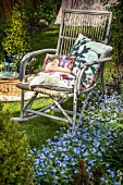 Weathered, comfortable cane rocking chair next to bed of forget-me-not in summery garden