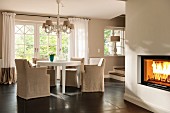 Pale, loose-covered chairs at round white table below chandelier: fire in fireplace to one side