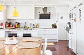 Place mats on wooden table below yellow pendant lamps in white fitted kitchen