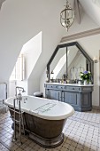 Free-standing vintage-style bathtub with floor-mounted taps on tiled floor and custom washstand with grey base cabinet in attic bathroom