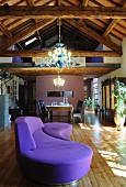 Curved purple sofa in loft apartment with wooden roof structure, lit chandeliers and dining area below gallery in background