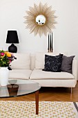 Glass coffee table in front of white sofa below sunburst mirror on wall
