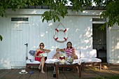 Two women and toddler on outdoor easy chairs below life belt on wall of white changing hut