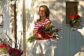 Fröhliche Frau mit roten Gladiolen im Arm vor weißem Holzhäuschen