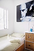Corner of a room with a single bed in front of a window with blinds, bedside table and portrait poster