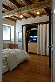 Doppelbett und begehbarer Kleiderschrank und Fernseher hinter Vorhang im Schlafzimmer mit rustikaler Holzbalkendecke