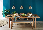Laid dining table with wooden bench under pendant lights in front of petrol-colored wooden paneling