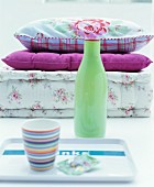Pale green vase between striped beaker on tray and stacked cushions with vintage floral patterns