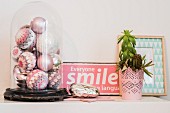 Various pastel Christmas baubles with crocheted covers under vintage glass cover next to pink enamel sign and plant on surface