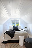 Double bed with pale and dark blankets below window in attic room with white wood-clad walls