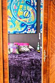 Bed with purple fur blanket and colourful artwork reflected in mirror on wardrobe door