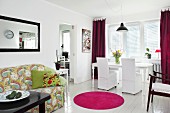 Offener Wohnbereich mit weißen Hussenstühlen und Esstisch auf weißem Dielenboden, pinkfarbener runder Teppichvorleger vor gemustertem Sofa