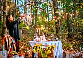 Woman decorating set table in autumnal woodland with fairy lights and crystal chandelier